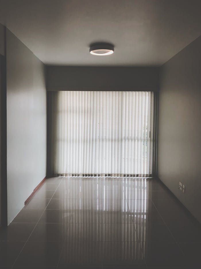about-us-01 Spacious empty room with modern design featuring large window blinds and tiled floor.